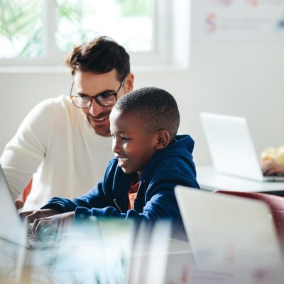 Male teacher helps a young boy with computer-based learning in a classroom setting. Child tutor providing a lesson in an elementary school, with a focus on coding and basic digital literacy.